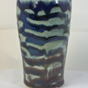 May include: A handmade ceramic tumbler with a unique glaze. The colors include dark blue, light blue, and brown, creating a layered, textured effect. The tumbler has a cylindrical shape and a slightly flared rim.