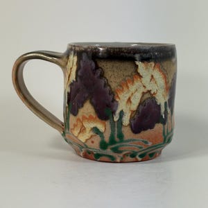 May include: A ceramic mug with a rounded handle, featuring a speckled tan base and a dark brown rim. The exterior is decorated with a repeating pattern of stylized purple and cream-colored floral shapes, accented by thin green stems at the bottom.