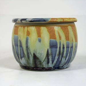 Op de afbeelding: Een keramische botervloot met een deksel, met een kleurrijk abstract ontwerp in tinten blauw, geel en oranje. Het ontwerp lijkt op een bloemenveld.