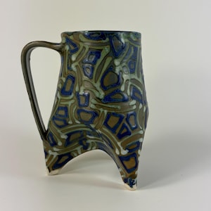 May include: A ceramic mug with a brown base color featuring a raised, textured pattern of blue and light green geometric shapes. The mug has a single handle on the left side and stands on three distinct, pointed feet at the base of the vessel.