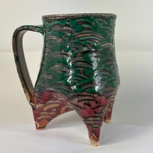 Puede incluir: Una taza de cerámica hecha a mano con un diseño único. La taza presenta una combinación de colores verde, rojo y beige con un patrón texturizado. Tiene un asa y está sostenida por tres patas. La taza es una obra de arte funcional.