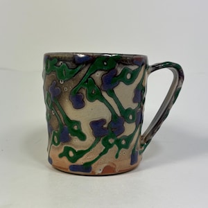 Op de afbeelding: Een handgemaakte keramische mok met een uniek bloemmotief. De mok heeft een crèmekleurige basis met groene, blauwe en bruine bloempatronen. De handgreep en de rand zijn ook versierd met groene en bruine accenten. Perfect voor warme dranken.