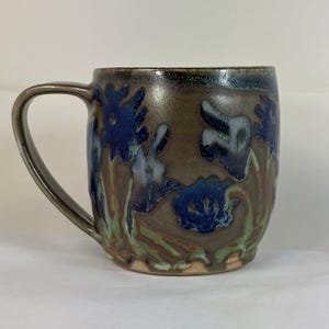 May include: Handmade ceramic mug with a dark brown base and blue floral design. The mug features a curved handle and is adorned with green and blue accents. A unique piece for enjoying your favorite beverage.
