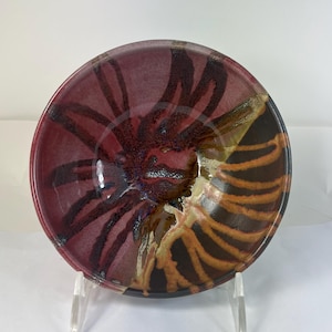 May include: A handmade ceramic bowl with a unique design. The bowl features a deep red interior with dark brown and black streaks radiating from the center. The exterior has a mix of brown, orange, and cream colors. The bowl is displayed on a clear stand.