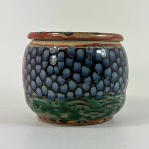 May include: A round ceramic jar with a lid featuring a glossy finish. The body displays a pattern of blue, rounded shapes resembling grapes or bubbles over a green base, with a reddish-brown rim and lid edge, creating a textured appearance.