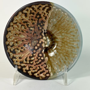 May include: A circular ceramic bowl featuring a dual-toned glaze. The left side displays a dark brown base with raised, textured, light tan branching patterns. The right side features a smooth, translucent, light blue and amber glaze.