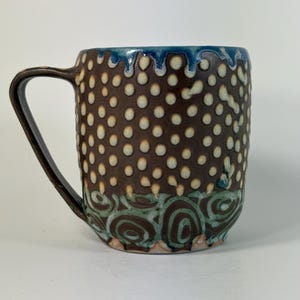 May include: A ceramic mug with a dark brown base color featuring a textured pattern of raised cream-colored dots across the upper body. The rim has a blue drip glaze, while the lower section displays a swirling green and brown pattern.