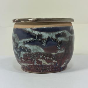 Op de afbeelding: Een handgemaakte keramische pot met een deksel. De pot heeft een unieke glazuur met een mix van donkerblauwe, bordeauxrode en lichtblauwe tinten. Het deksel is een gedempt grijsbruin. De pot heeft een ronde vorm.