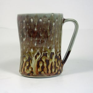 May include: A ceramic mug with a light green glaze and brown and white abstract pattern. The mug has a handle and a rounded shape.