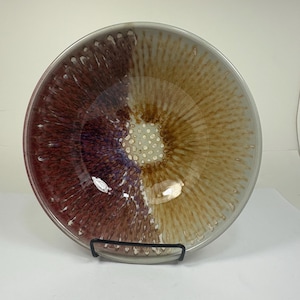 May include: A ceramic bowl with a unique color gradient. The bowl transitions from a deep burgundy on the left to a golden brown on the right, with a speckled center. The rim is a light gray. The bowl is on a black metal stand.