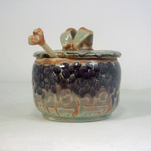 May include: A handcrafted ceramic jar with a lid and a small spoon. The jar is adorned with a textured pattern of dark purple and brown circles, with green and orange accents. The lid features a decorative handle. A unique kitchen or home decor item.