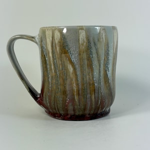 May include: A ceramic mug featuring a vertical drip glaze pattern in shades of grey, tan, and deep red. The mug has a smooth, cylindrical body with a curved handle on the left side. The glaze creates a textured, layered appearance across the surface.