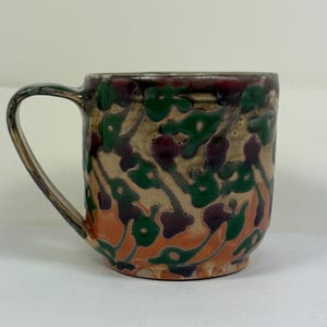 May include: A ceramic mug with a curved handle. The mug has a beige background with a repeating pattern of green and purple floral shapes, and orange accents near the base. The rim and handle have a dark brown glaze.