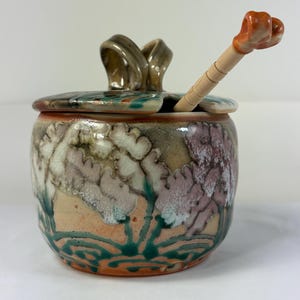 May include: A ceramic sugar bowl with a lid and spoon. The bowl is decorated with a floral design in shades of pink, green, and beige. The lid has a handle, and the spoon has an orange handle. The bowl is approximately 4 inches tall.