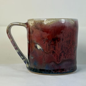 May include: A handmade ceramic mug with a unique glaze. The mug features a deep red color with streaks of black, blue, and cream. The handle is a matching color. This mug is perfect for coffee, tea, or any hot beverage.