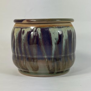 May include: A round ceramic jar with a lid, featuring a glossy glaze in shades of deep purple, muted green, and earthy brown. The surface displays a vertical drip pattern where the colors blend and overlap, creating a textured appearance.