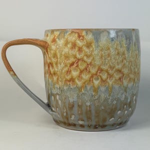 May include: A handmade ceramic mug with a unique textured glaze. The mug features a warm color palette of orange, yellow, and gray, with a brown handle. The design has a dripped effect, adding to its artisanal charm. Perfect for coffee or tea.