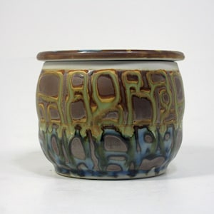 Op de afbeelding: Een keramische botervloot met een deksel. De vloot is versierd met een bruine, groene en blauwe glazuur. De glazuur heeft een gevlekt, abstract patroon.