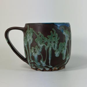 May include: A ceramic mug with a dark brown base color featuring a decorative drip glaze pattern in shades of green and light blue. The mug has a curved handle on the left side and a smooth, rounded body shape with a wide rim at the top.