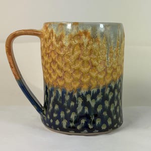 May include: A handmade ceramic mug with a textured surface. The mug features a gradient of colors, including yellow, blue, and brown. The handle is brown and blue. The mug is designed for hot beverages.
