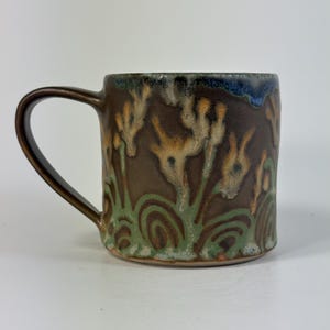 May include: A ceramic mug with a dark brown base color featuring a hand-painted pattern of green swirling lines at the bottom and light tan, flower-like shapes extending upward. The rim has a dark blue glaze accent and a curved handle.