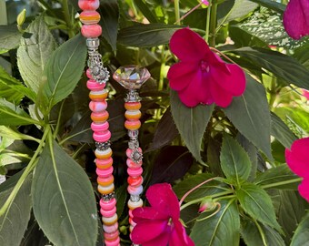 May include: Two decorative garden stakes with colorful beads and crystal-like toppers. The stakes feature a variety of pink, orange, and white beads, accented with silver-colored metal charms. The stakes are placed among green foliage and vibrant pink flowers.
