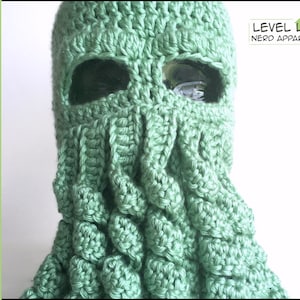 May include: A light green crochet balaclava with eye openings and textured details. The balaclava has a tentacle-like design around the neck and face. The text "CROCHET PATTERN" is on the left side, and "LEVEL UP NERD APPAREL" is on the right.