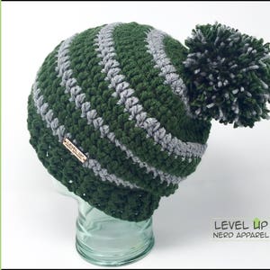 Könnte beinhalten: Grün-graue gestreifte Häkelmütze mit einem flauschigen Bommel oben. Die Mütze hat ein kleines Holzschild mit der Aufschrift "Level Up Nerd Apparel".