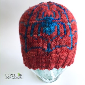 May include: Red knit beanie with a blue and red pattern. The beanie is on a glass display stand.