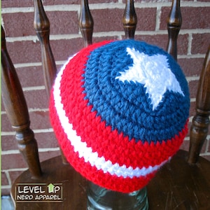 May include: A red, white, and blue crocheted hat with a white star on a blue background. The hat is sitting on a wooden chair.
