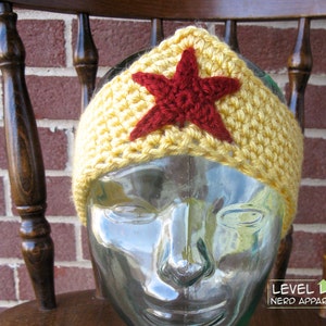 May include: Crocheted headband with a yellow base and a red star in the center. The headband is on a glass mannequin head. The text "LEVEL UP NERD APPAREL" is visible in the lower right corner of the image.