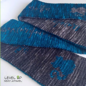 May include: Three knitted neck warmers in a teal and gray color scheme. The neck warmers have a repeating pattern of a stylized image of a dragon.