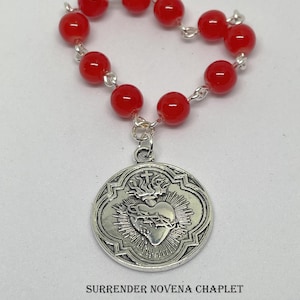 May include: Red glass bead rosary with a silver charm featuring a heart with thorns and a crown. The charm is inscribed with "SURRENDER NOVENA CHAPLET".