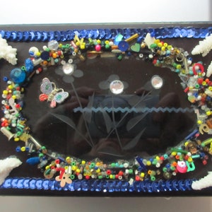 May include: A small, black, rectangular box with a glass top decorated with colorful beads, sequins, and a butterfly charm. The glass top has a design of flowers and a blue wavy line.