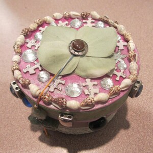 May include: A pink and white round box with a decorative flower made of fabric and a brown stone in the center. The box is decorated with seashells, rhinestones, and puzzle pieces.