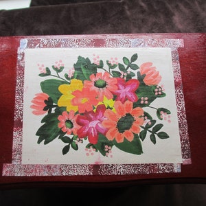 May include: A wooden box with a painted floral design. The box is painted red with a white border and a white center panel. The floral design features pink, red, orange, yellow, and green flowers.