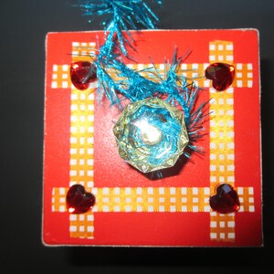 Może przedstawiać: Czerwony kwadrat z biało-pomarańczowym wzorem w kratkę. W środku znajduje się niebieski i zielony klejnot, otoczony trzema czerwonymi klejnotami w kształcie serca.