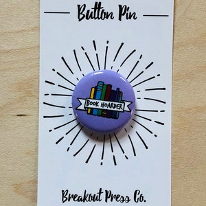 May include: Purple button pin with a stack of colorful books and a banner that reads "Book Hoarder". The pin is on a white card with the text "Button Pin" and "Breakout Press Co."