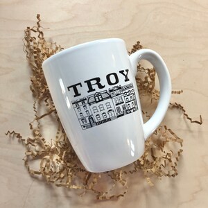 May include: White ceramic mug with a black graphic of a row of houses and the word "TROY" in black lettering.