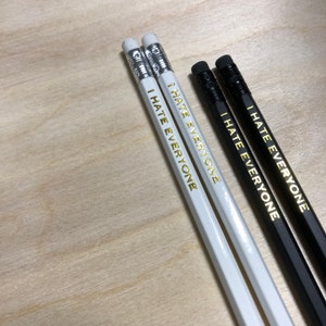 May include: Four pencils, two white and two black, with silver erasers. The pencils are printed with the phrase "I HATE EVERYONE" in gold lettering. The pencils are arranged on a light wood surface.