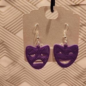 Op de afbeelding: Een paar paarse oorhangers met een ontwerp van een vrolijk en een verdrietig gezichtsmasker. De oorhangers zijn gemaakt van een getextureerd materiaal en hebben zilveren haakjes.