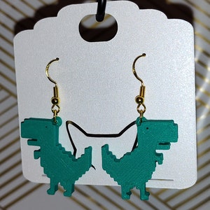 May include: A pair of teal-colored dinosaur earrings with gold-colored ear wires. The earrings feature a pixelated design, giving them a retro video game aesthetic. The earrings are displayed on a white card.