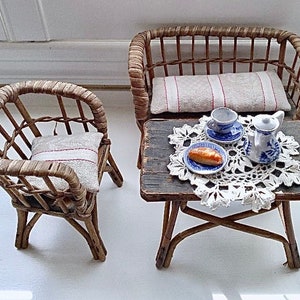 May include: Miniature wicker chairs with cushions, arranged around a small wooden table. The table is set with a blue and white tea set, a plate with a pastry, and a white lace doily. A charming, rustic scene.