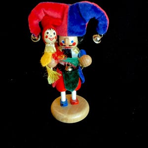 May include: A wooden nutcracker figurine dressed as a jester in a red and blue velvet hat with silver bells. The figure holds a small doll with a yellow tassel, wears a green and red tunic with a gold collar, and stands on a round wooden base.