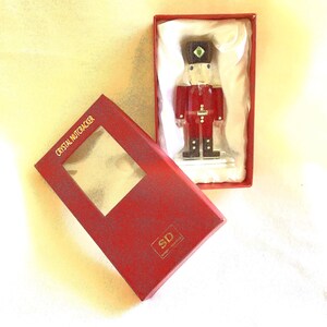May include: A clear crystal nutcracker figurine with red accents, a black hat, and a green gem detail, presented in a red box with a clear window. The box lid features the text CRYSTAL NUTCRACKER and the initials SD in gold lettering.