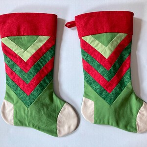 Könnte beinhalten: Zwei Weihnachtsstrümpfe mit rotem Umschlag und einem Chevron-Muster aus abwechselnden hellgrünen, dunkelgrünen und roten Stoffstreifen. Der untere Teil ist einfarbig grün mit cremefarbener Spitze und Ferse sowie einer roten Aufhängeschlaufe.