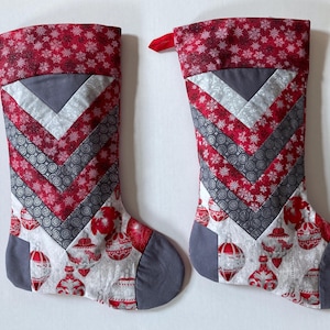May include: Two Christmas stockings featuring a chevron pattern of red snowflake fabric, grey fabric, and silver patterned fabric. The lower sections display a white background with red and silver ornament prints and solid grey heels and toes.