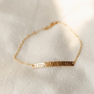 May include: A gold-coloured bracelet featuring a rectangular bar pendant with an engraved floral design. The bracelet is shown on a white, textured surface. The delicate chain and pendant create an elegant accessory.