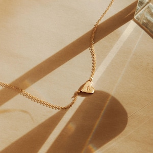 May include: A gold-coloured necklace featuring a heart-shaped pendant. The pendant is engraved with the letter "H". The necklace rests on a light brown surface, with shadows across it.