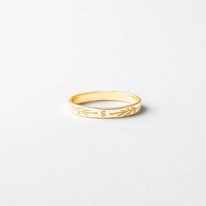 Puede incluir: Un anillo dorado con un diseño de hojas grabado y la letra "S". El anillo está sobre un fondo blanco. Es una banda delicada, probablemente para uso diario.